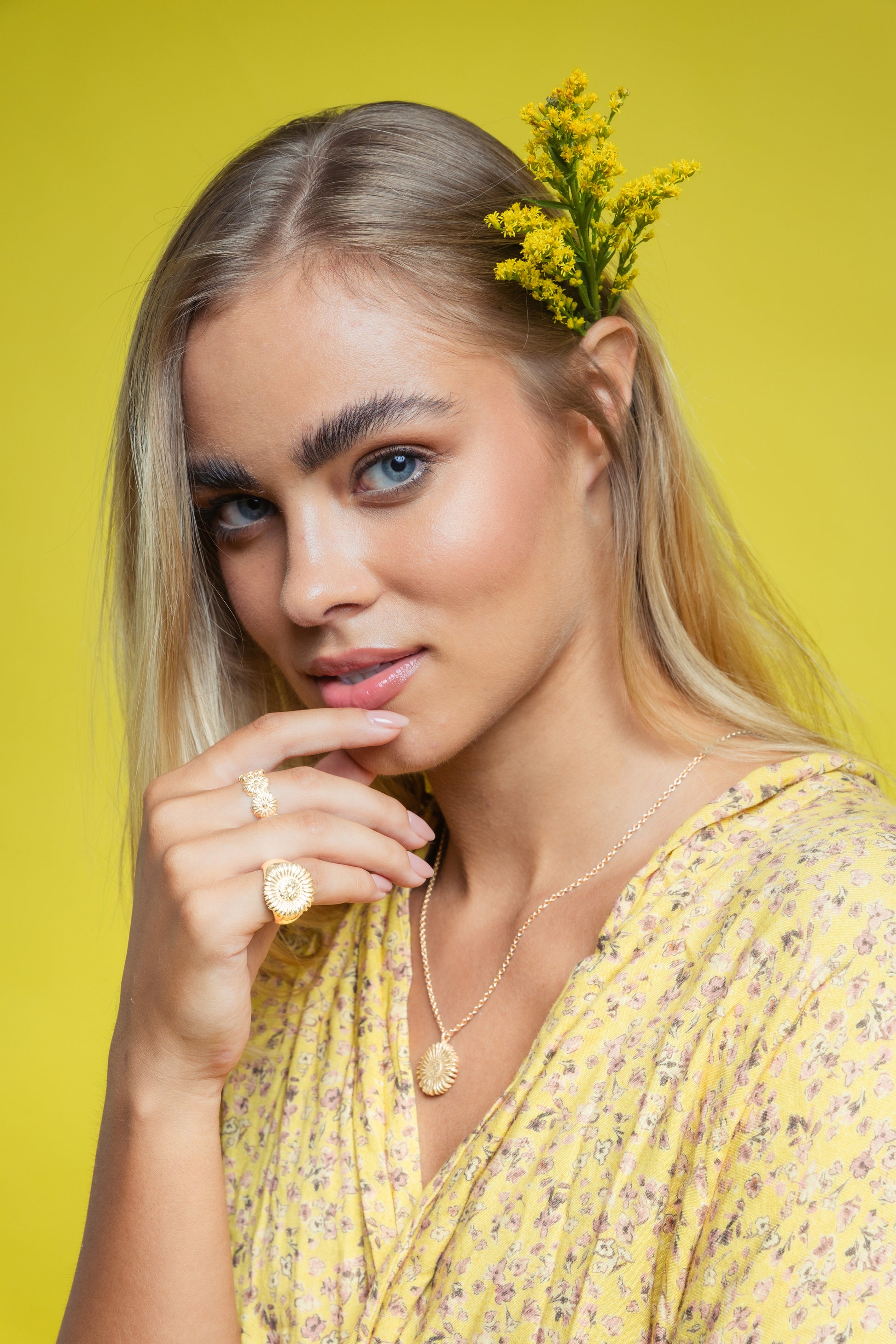 Blonde girl on yellow background , wearing a yellow dress with a yellow flower tucked into her left ear. She has a gold sunflower necklace and wears two gold sunflower, handmade by Saint Davis rings on her right hand. 