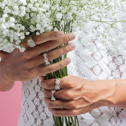 Close up of mans hands holding small bouquet of white flowers. He has 4 silver handmade sunflower rings on handmade by Saint Davis.