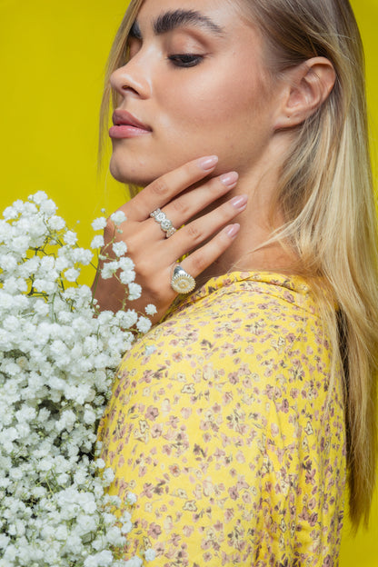 Blonde girl turned to the side, holding her hand to her face, her right hand has two silver floral, handmade rings by Saint Davis. She is wearing a yellow dress against a yellow background and holding a small bouquet of white flowers. 